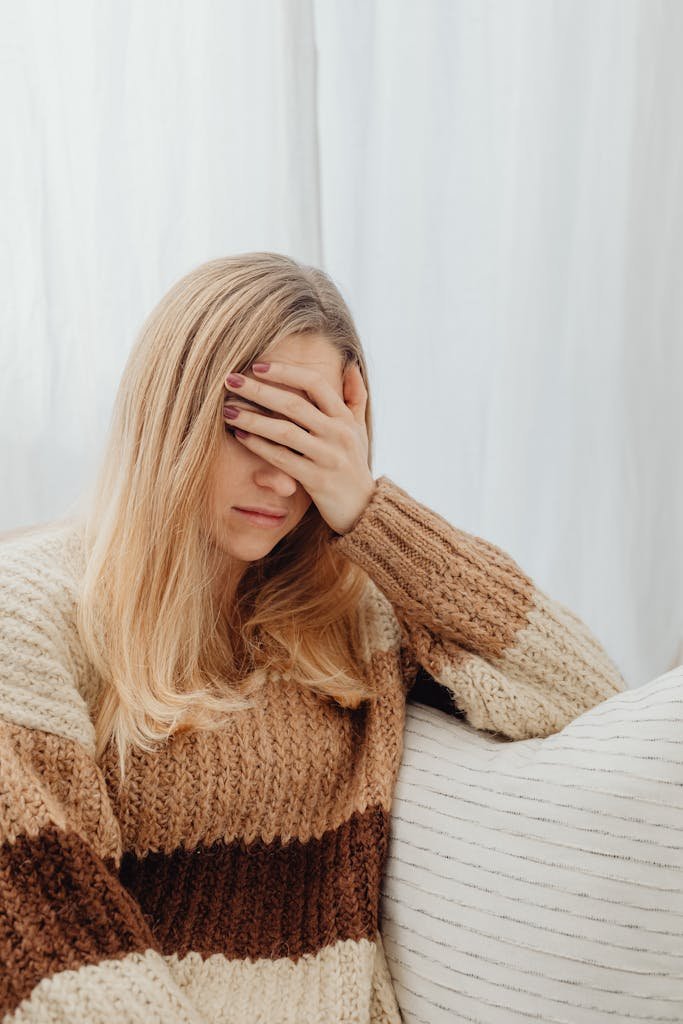 A woman in a warm sweater covers her face, appearing unwell while sitting indoors.