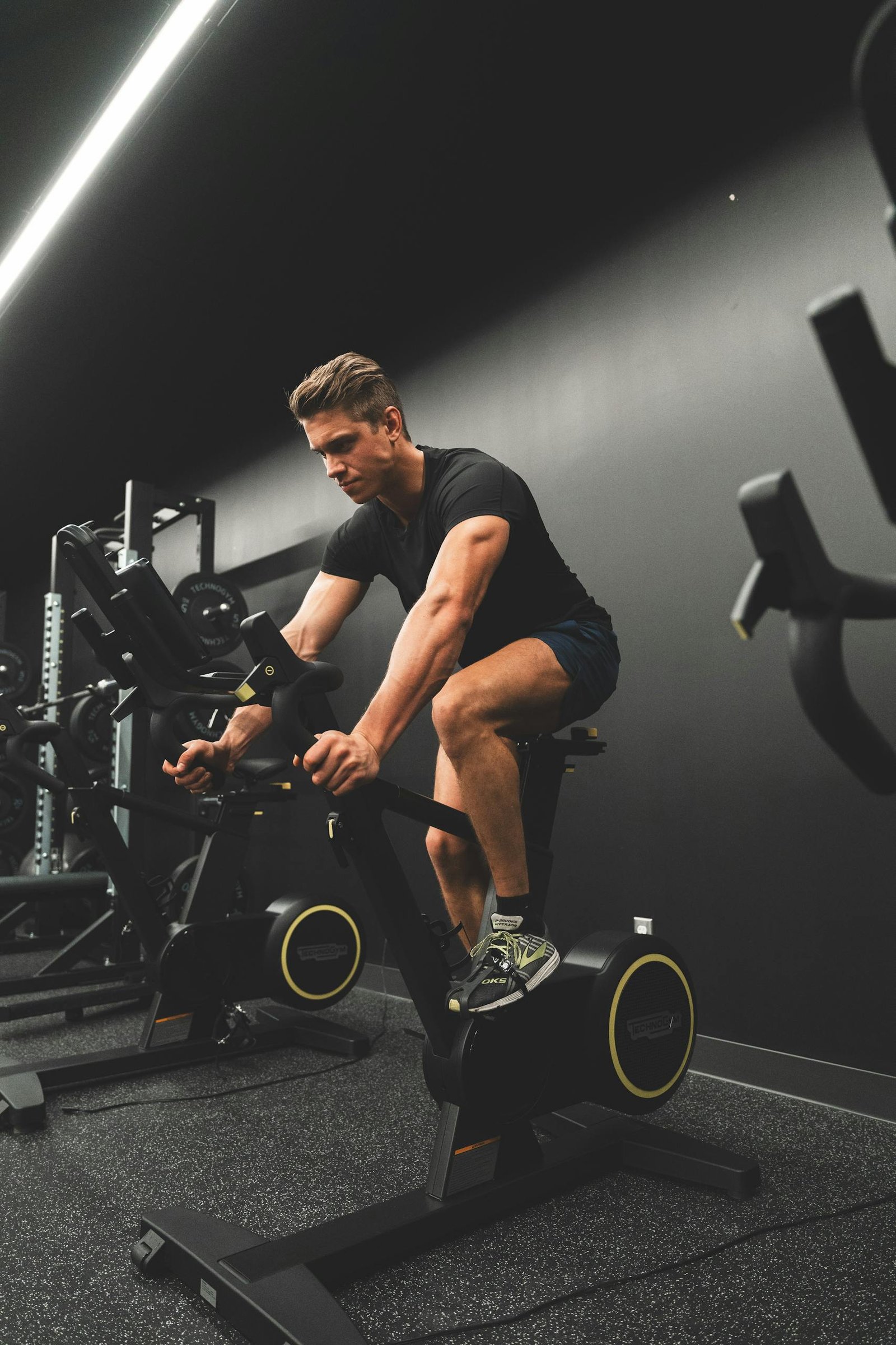 Muscular man biking indoors in gym setting, showcasing fitness and sportswear.